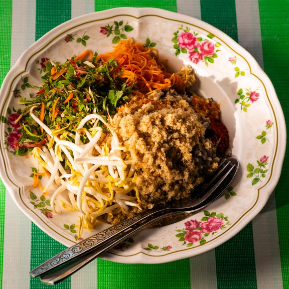 In this salad, rice gets topped with pinches of toasted coconut, dried fish, finely-sliced herbs and vegetables, bean sprouts, a tiny knot of noodles, crushed black pepper and dried chili. 