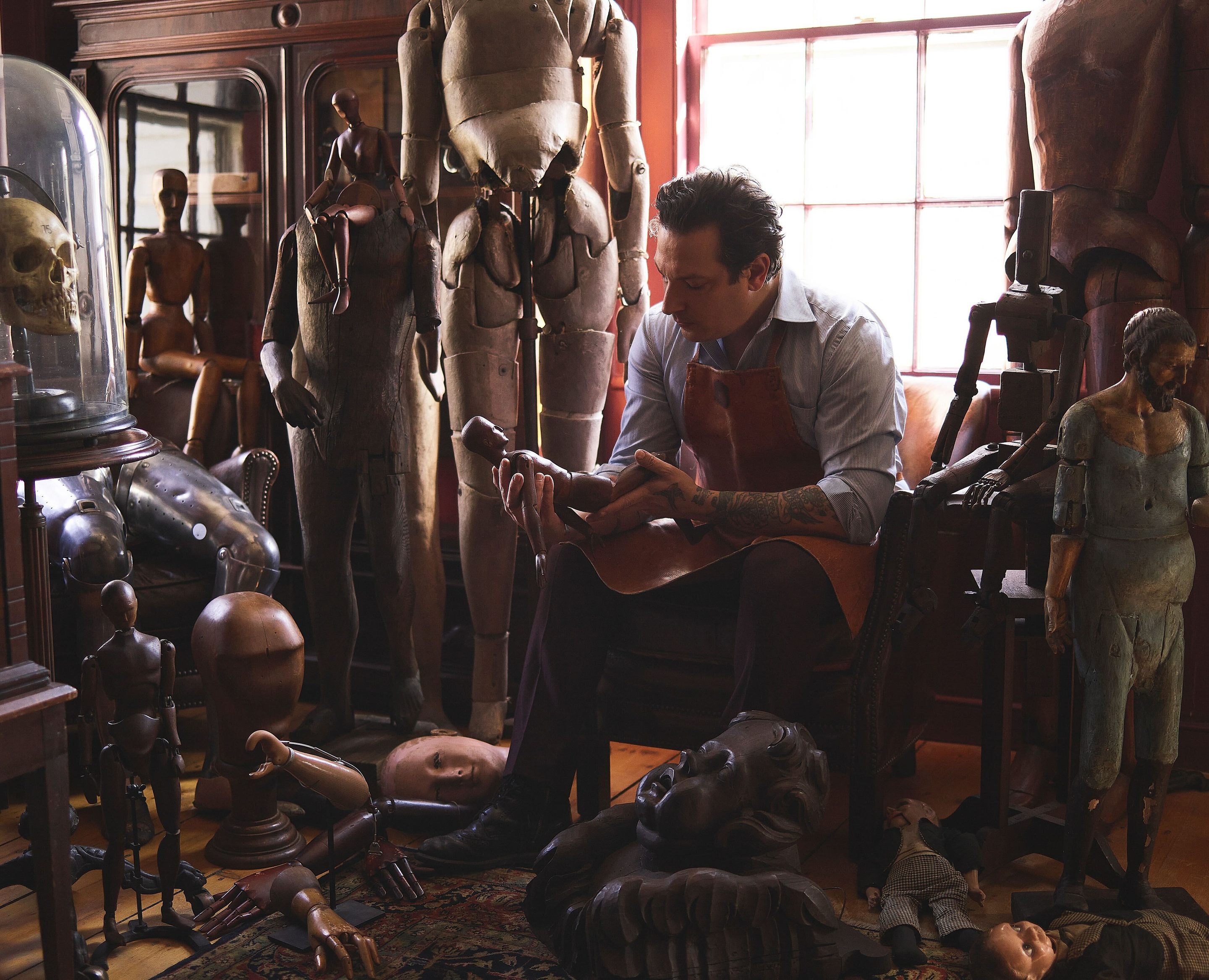 A man sitting in a workshop, surrounded by wooden figure sculptures, looks at a doll in his hands.